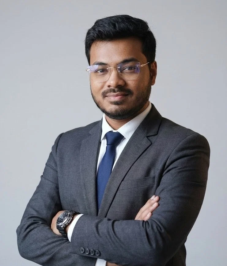 WhatsApp-Image-2025-12-24-at-7.06.14-AM - Kihan Marketing - Kihan Marketing Professional headshot of a man in a dark gray suit, white shirt, and blue tie, with arms crossed against a neutral gray background, wearing glasses and a slight smile.