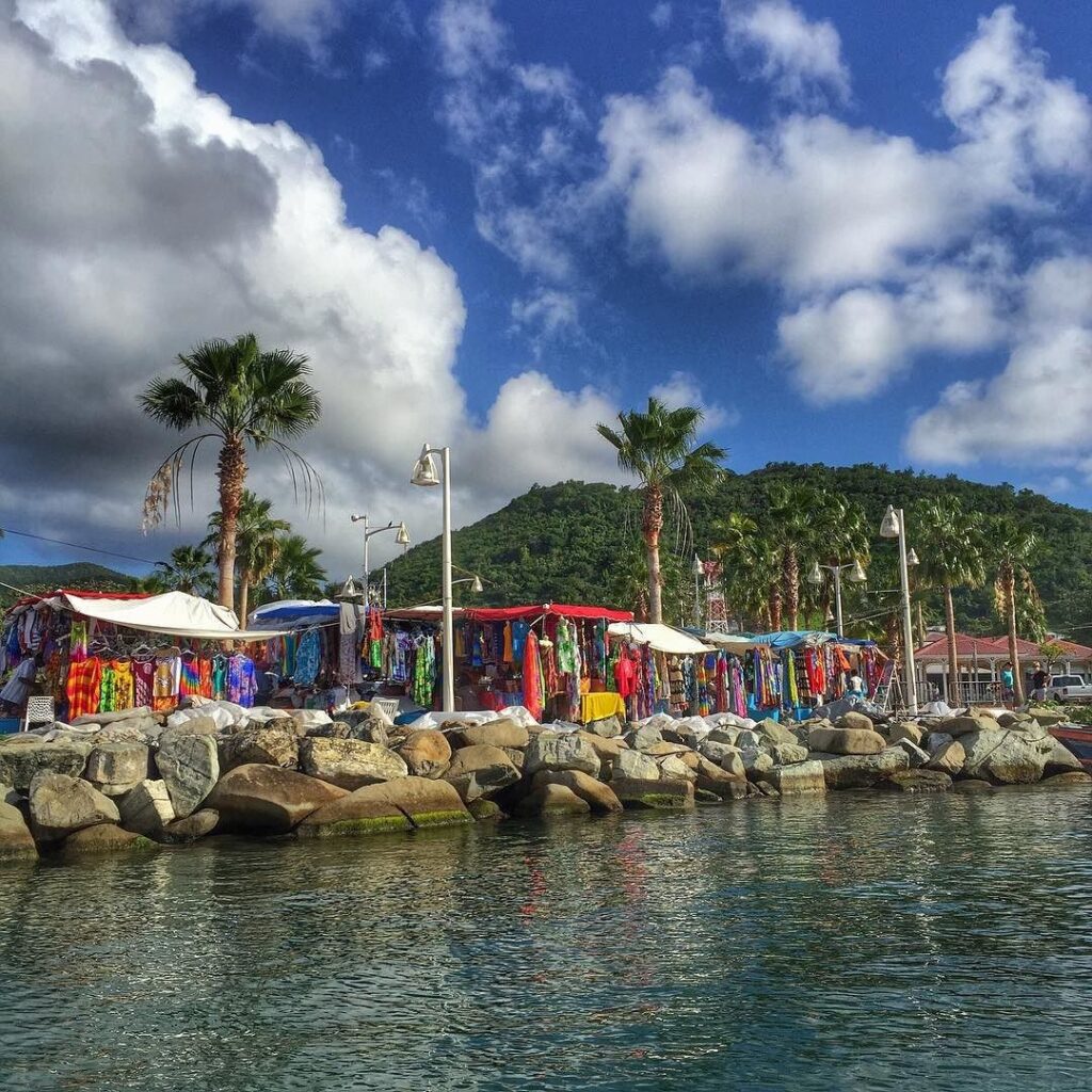 a063c1adc169b8a9460b8fd67fbee729 - Kihan Marketing - Kihan Marketing Colorful seaside market along a rocky jetty with clothing stalls, palm trees, and a blue sky over green hills.