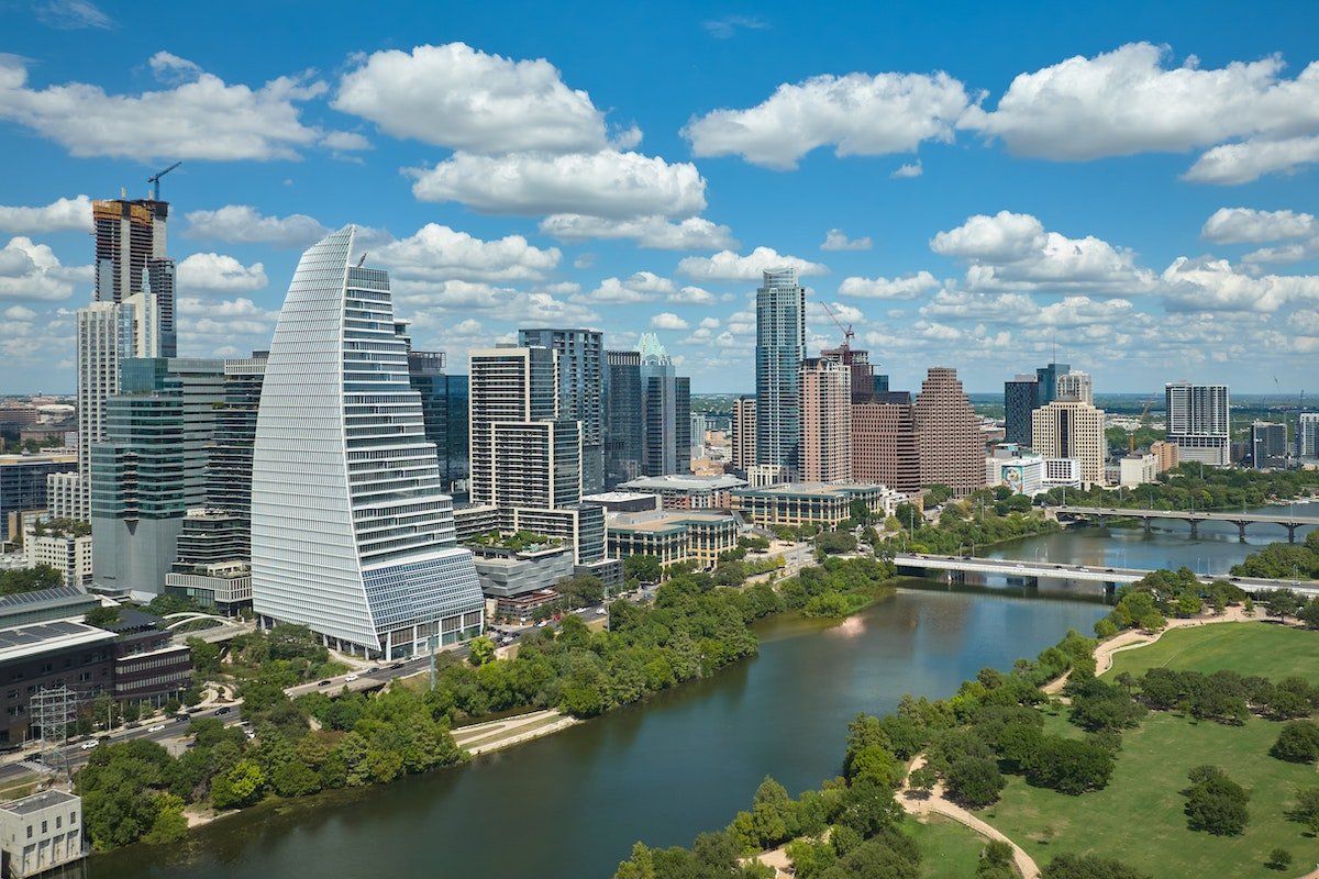 Aerial view of downtown Austin, Texas along the Colorado River with a distinctive curved white skyscraper and modern glass buildings under a blue sky with clouds.