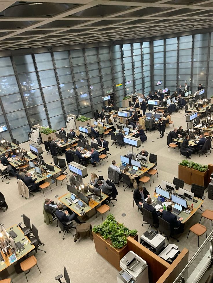 Open-plan office trading floor with rows of desks, monitors, and people at computers near large glass windows.