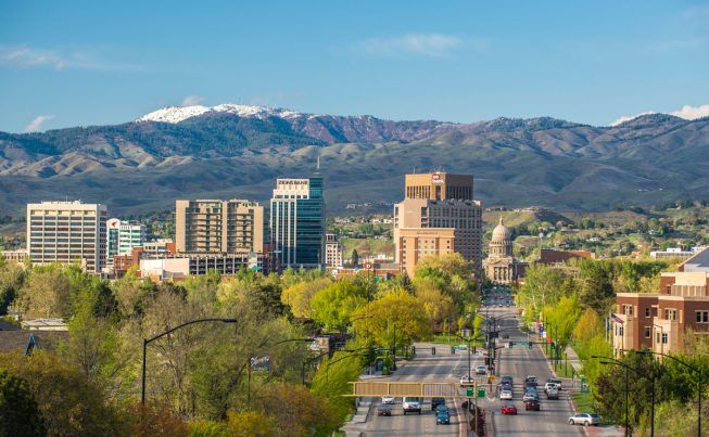 City skyline with modern high-rises, tree-lined street in foreground, and snow-capped mountains in the distance.