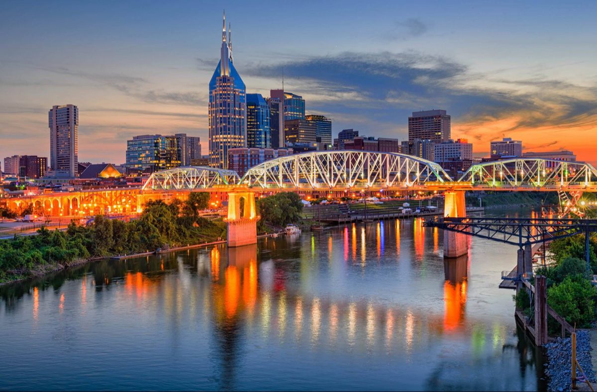 Dusk city skyline with a brightly lit steel truss bridge spanning a calm river and colorful reflections on the water.