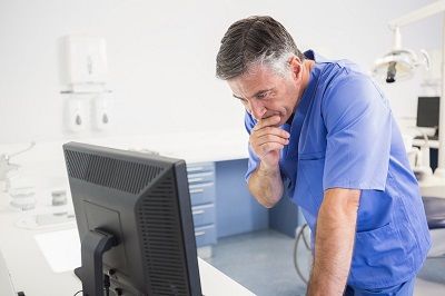 Male healthcare professional in blue scrubs leaning over a computer monitor in a clinical room, thinking with a finger on his chin.