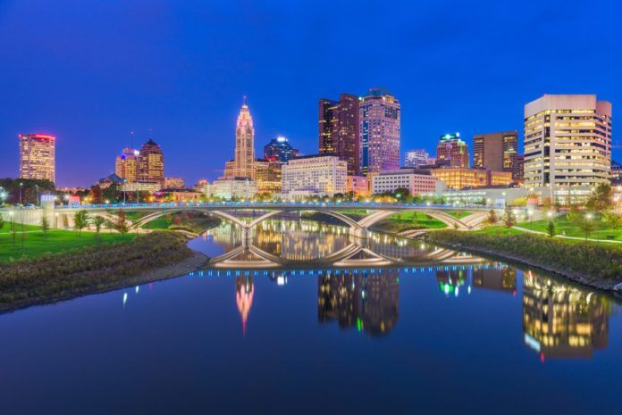 Night city skyline over a calm river, with a lit arched bridge and its reflection in the water.