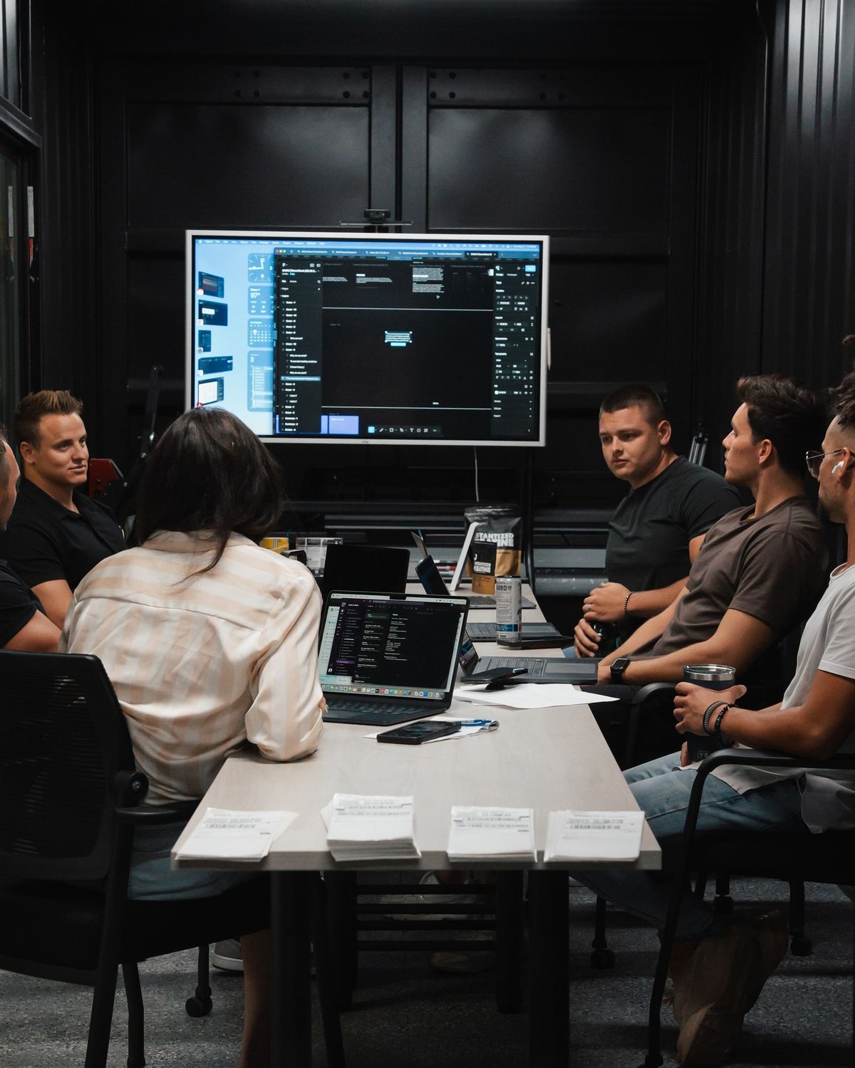 Six people seated around a conference table, facing a large monitor displaying a dark code/editor UI with laptops and papers on the table.