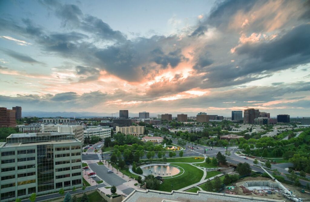 c9035f68d806a2b7dcf00749d3ff5f88 - Kihan Marketing - Kihan Marketing Panoramic view of a city skyline at sunset with a circular fountain and green park in the foreground, roads and buildings lining the area.