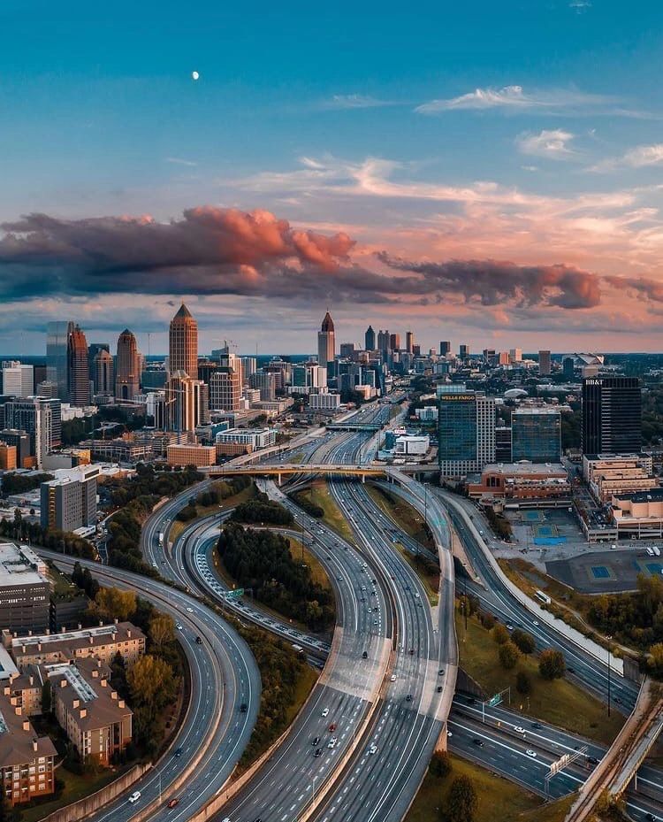c98ab5e36a4026a8996849ed2cb5144e - Kihan Marketing - Kihan Marketing Aerial view of a city with winding elevated highways and a skyline at sunset. Cars travel on multiple freeway loops beneath tall buildings.