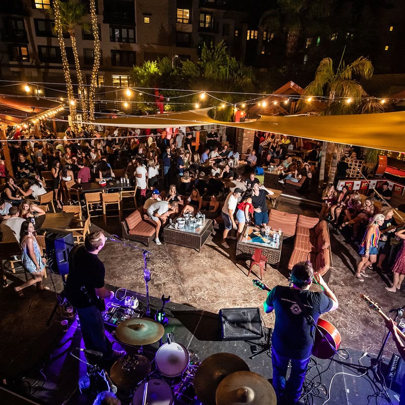 Outdoor nighttime party with a live band performing on stage while a crowd mingles under string lights and tents.