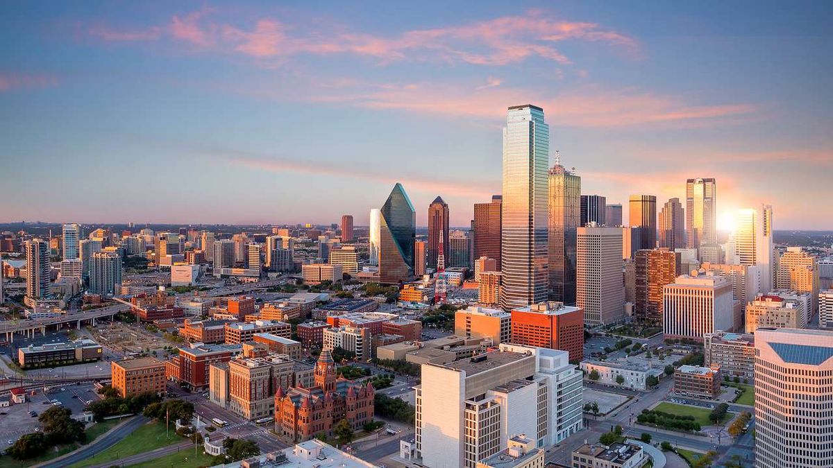Aerial view of a downtown skyline at sunset with tall glass skyscrapers and a warm orange-pink sky.
