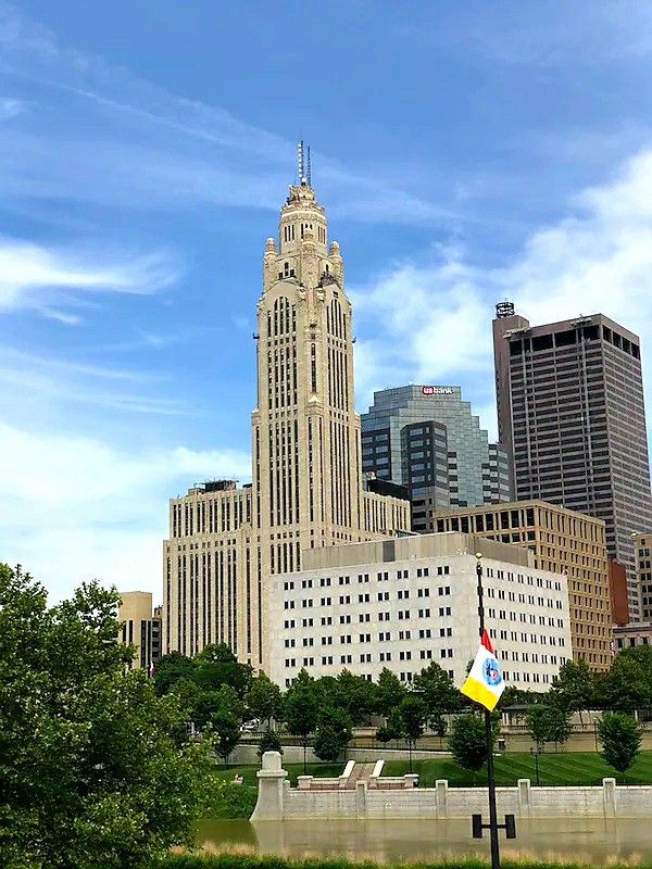 f8a3e19bd9eee47b2f54fe71e9472e66 - Kihan Marketing - Kihan Marketing Tall beige art-deco tower rises above city skyscrapers; trees and a river in the foreground with a flag on a pole.