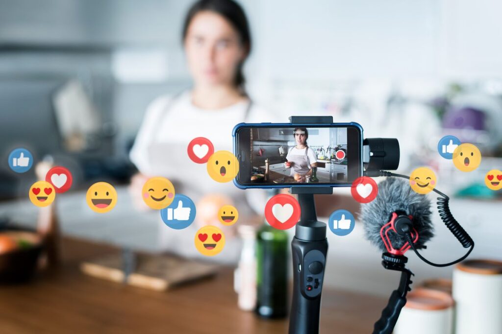 Content creator filming a cooking video with a phone on a stabilizer, while floating social emojis indicate engagement.