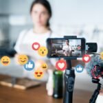 7d7fa7f331dbf907a636cc6c2f173f7e - Kihan Marketing Content creator filming a cooking video with a phone on a stabilizer, while floating social emojis indicate engagement.
