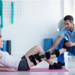Screenshot - Kihan Marketing Physical therapist assists a woman with knee braces during a leg-raise exercise on a pink mat in a rehab setting.