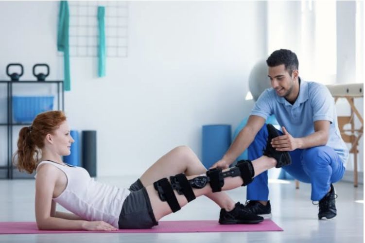 Physical therapist assists a woman with knee braces during a leg-raise exercise on a pink mat in a rehab setting.