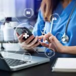 Healthcare worker in blue scrubs uses a smartphone with medical icons and a laptop on the desk while a stethoscope rests nearby.