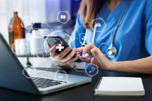 Healthcare worker in blue scrubs uses a smartphone with medical icons and a laptop on the desk while a stethoscope rests nearby.