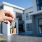de72d9cb90a4fdc0b6f6b05dc2610d15 - Kihan Marketing Person holding a key with a wooden house-shaped keychain in front of a modern house, symbolizing home ownership or moving in.