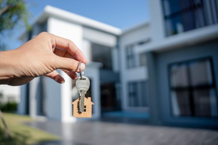 Person holding a key with a wooden house-shaped keychain in front of a modern house, symbolizing home ownership or moving in.