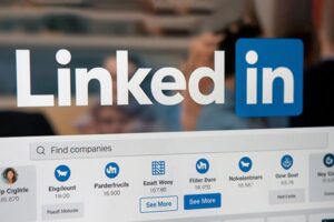 LinkedIn logo with a blurred workspace on the screen showing a Find companies search bar and 'See More' buttons beneath company icons.