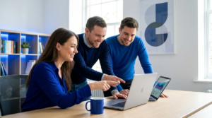 Team of three colleagues collaborates around a desk, one woman points at a laptop while two men look on, smiling in a bright office.