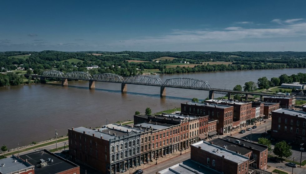 Aerial view of a wide river with a metal truss bridge spanning it, and a row of red-brick storefront buildings along the riverside town.