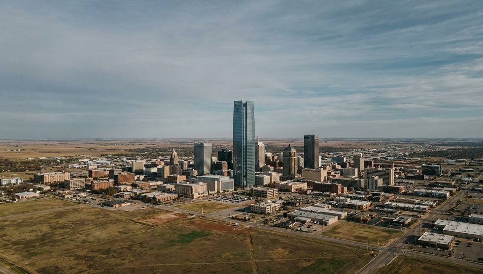 Aerial view of a city skyline with two tall glass skyscrapers surrounded by low-rise buildings on a clear day, with open fields nearby.