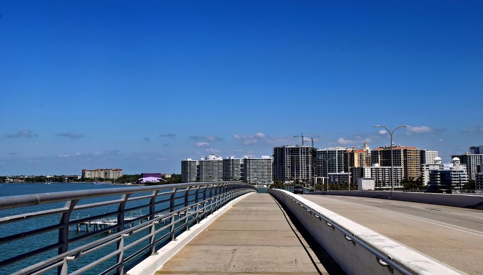 Pedestrian bridge walkway over calm water toward a city skyline of high-rise buildings under a clear blue sky.